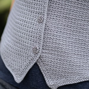 Op de afbeelding: Close-up van een lichtgrijs gehaakt vest met drie decoratieve zilveren knopen. Het vest heeft een textuur, open geweven ontwerp. De knopen zijn rond met een ingewikkeld bloemenpatroon. Het vest wordt gedragen over een donkerblauw kledingstuk.
