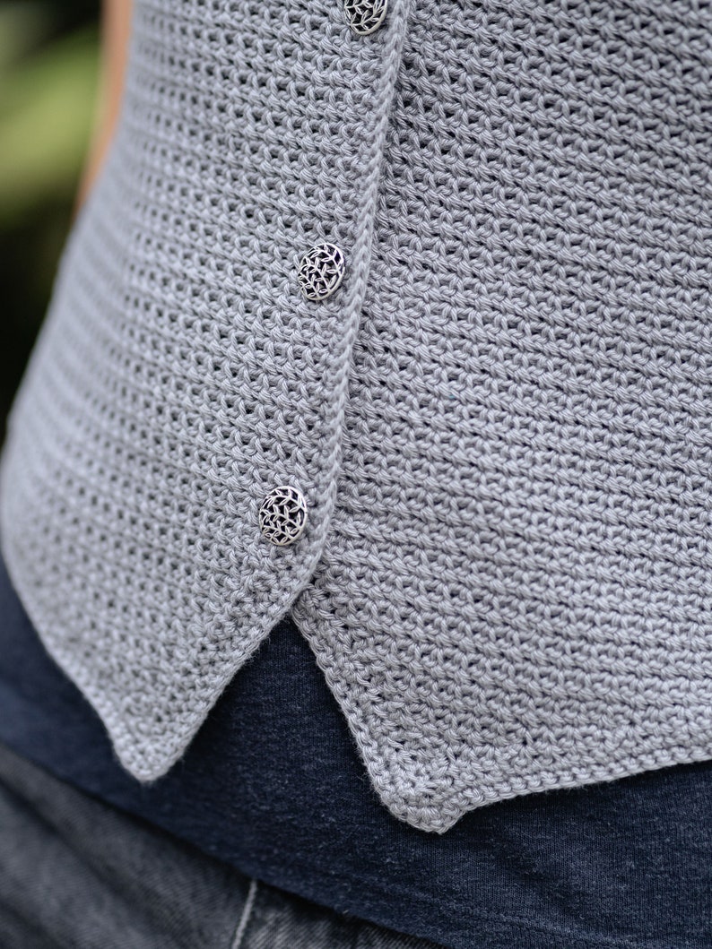 Op de afbeelding: Close-up van een lichtgrijs gehaakt vest met drie decoratieve zilveren knopen. Het vest heeft een textuur, open geweven ontwerp. De knopen zijn rond met een ingewikkeld bloemenpatroon. Het vest wordt gedragen over een donkerblauw kledingstuk.