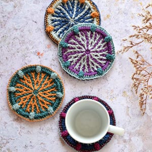 May include: Four crocheted coasters in different colours and patterns. The coasters are round and have a beaded border. A white ceramic mug is sitting on one of the coasters.
