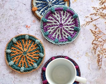 May include: Four crocheted coasters in different colors and patterns. The coasters are round and have a beaded border. A white ceramic mug is sitting on one of the coasters.