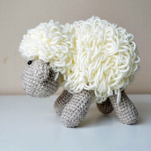 Puede incluir: Un juguete de oveja de crochet blanco con un cuerpo marr&oacute;n y ojos negros. La oveja est&aacute; de pie sobre una superficie blanca.
