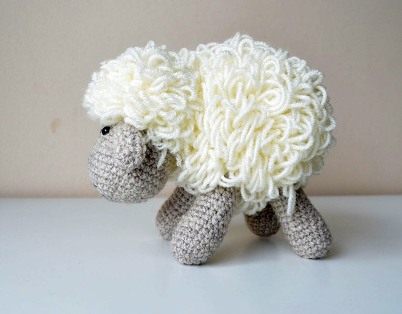 Puede incluir: Un juguete de oveja de crochet blanco con un cuerpo marr&oacute;n y ojos negros. La oveja est&aacute; de pie sobre una superficie blanca.