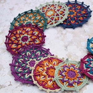 May include: A set of colourful crocheted coasters with intricate patterns. The coasters are made in shades of purple, teal, orange, and cream.
