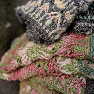 May include: A folded crochet blanket featuring intricate floral patterns in shades of charcoal gray, cream, sage green, and dusty rose. The textile displays a dense, textured stitch construction with repeating geometric and botanical motifs throughout.