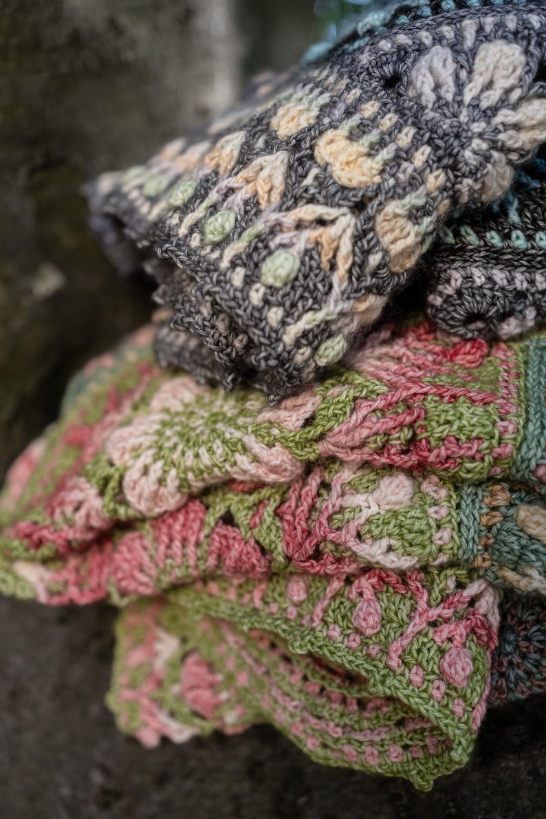 May include: A folded crochet blanket featuring intricate floral patterns in shades of charcoal gray, cream, sage green, and dusty rose. The textile displays a dense, textured stitch construction with repeating geometric and botanical motifs throughout.