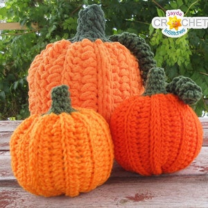 May include: Three crocheted pumpkins in various sizes, the largest is orange with a green stem, the medium pumpkin is orange with a green stem, and the smallest pumpkin is orange with a green stem. The pumpkins are sitting on a wooden table.