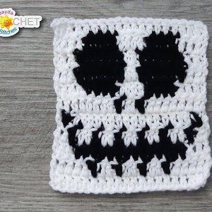 May include: A white crocheted square with a black crocheted Jack Skellington face design. The square is on a gray wooden surface.