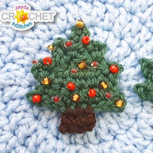 May include: Two green crochet Christmas tree ornaments with brown trunks. One tree is decorated with red beads and gold accents. The background is a light blue crocheted fabric. The logo "Jayda Crochet InStitches" is in the top left corner.
