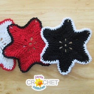 May include: Three crocheted dishcloths in the shape of stars. One is white with a red border, one is red with a black border, and one is black with a white border. The dishcloths are on a wooden surface.
