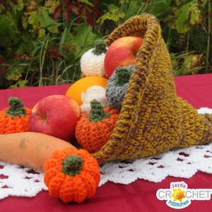 May include: A crocheted cornucopia filled with crocheted pumpkins, apples, and a sweet potato. The cornucopia is brown and gold and is sitting on a white crocheted doily on a red table. The text "Jayda Crochet Stitches" is visible in the lower right corner of the image.