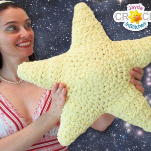 May include: A woman holds a large, yellow crocheted star-shaped pillow in front of a dark blue background with white stars. The pillow is soft and fluffy, and the woman is smiling.