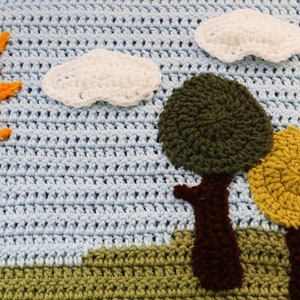 May include: A crocheted baby blanket with a blue background, a yellow sun, white clouds, and three trees with brown trunks. The blanket has a green and brown border.