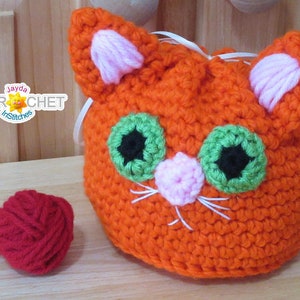 Può includere: Borsa a cordoncino a forma di gatto di colore arancione realizzata a uncinetto, con occhi verdi, naso e orecchie rosa e baffi bianchi. In primo piano c'è un gomitolo di lana rosso.
