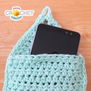 Puede incluir: Un soporte para teléfono de crochet azul claro con un teléfono inteligente negro en su interior. El soporte tiene un lazo en la parte superior para colgarlo.