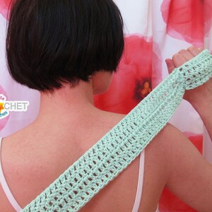 May include: A light green crocheted back scrubber with a textured pattern. The scrubber is being held by a person in front of a white shower curtain with red poppy flower designs.