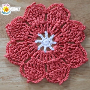 May include: A coral crochet flower with a white crochet center. The flower is on a wooden surface.
