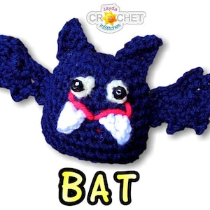 May include: A crocheted blue bat with white teeth and red mouth. The bat has large, black eyes and is flying with its wings spread out. The word "BAT" is written in yellow letters below the bat.