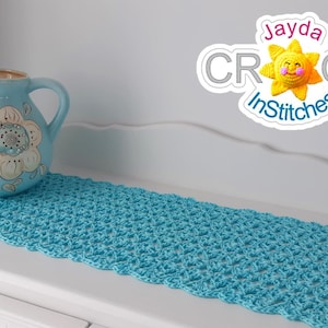 Può includere: Un runner da tavolo all'uncinetto blu chiaro con un bordo a festoni. Il runner è su una superficie bianca con un'anfora di fiori blu e bianca.