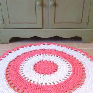 May include: A handmade, circular area rug with a crochet pattern. The rug features concentric circles of white and coral pink. The rug is on a tiled floor in front of a light green cabinet. Text on the image reads "Beautiful Full Circle Area Rug Pattern."