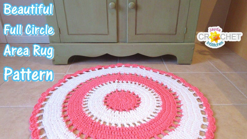 May include: A handmade, circular area rug with a crochet pattern. The rug features concentric circles of white and coral pink. The rug is on a tiled floor in front of a light green cabinet. Text on the image reads "Beautiful Full Circle Area Rug Pattern."