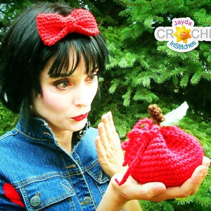 May include: A woman wearing a blue denim jacket and a red bow headband holds a red crocheted apple in her hand. The apple has a small brown stem and a white leaf. The background is a green leafy tree.