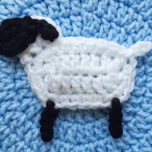 Op de afbeelding: Een gehaakte schaap applicatie met een witte body, zwarte kop en poten en een kleine staart. Het schaap staat tegen een getextureerde lichtblauwe achtergrond. Het logo "Jayda Crochet InStitches" staat in de linkerbovenhoek.