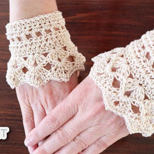 Könnte beinhalten: Ein Paar gehäkelte Armbänder in Cremefarbe. Die Armbänder haben ein zartes Spitzenmuster und werden an den Handgelenken einer Person gezeigt.