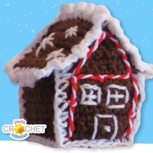 May include: A crocheted gingerbread house ornament with white icing and red candy canes. The house is brown and has white windows and a door. The ornament is against a blue background with white snowflakes.