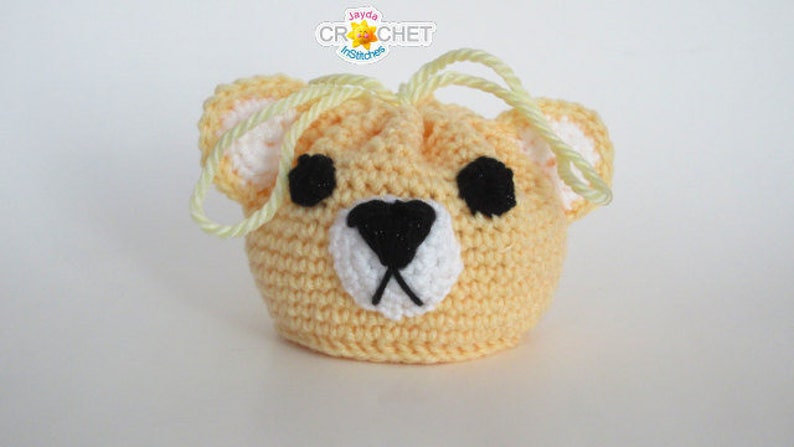 May include: A crocheted drawstring bag shaped like a bear's face. The bag is yellow with black eyes, a black nose, and a white mouth. The drawstring is yellow.