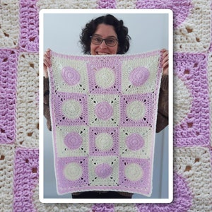 May include: A crocheted baby blanket with a white and purple square pattern. The blanket is held by a person in the center of the image. The text "Jayda Crochet In Stitches" is in the top left corner of the image.