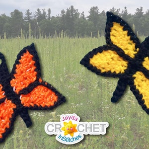 May include: Two crocheted butterflies, one orange and one yellow, with black outlines, are displayed against a blurred background of a field of tall grass.