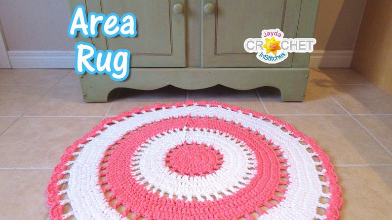 May include: A handmade, circular area rug in a spiral design. The rug features alternating bands of white and coral pink, with a scalloped edge. The words "Area Rug" are in a blue bubble font. The rug is placed on a tiled floor.