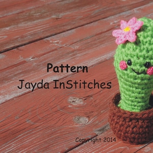 May include: A green crocheted cactus with a pink flower on top, a smiling face, and brown pot. The cactus is sitting on a wooden surface. The text "Pattern Jayda InStitches Copyright 2014" is visible below the cactus.