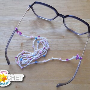 May include: White crocheted eyeglass chain with pink and blue beads. The chain is attached to a pair of black eyeglasses with gold accents. The chain is a handmade item and is perfect for keeping your glasses from falling off.