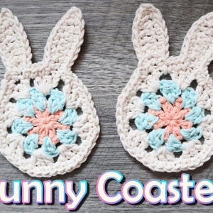 May include: Two crocheted bunny-shaped coasters with a pink and blue flower design in the center. The coasters are on a gray wooden surface.  "Bunny Coasters" is written in pink, blue, and white letters below the coasters.