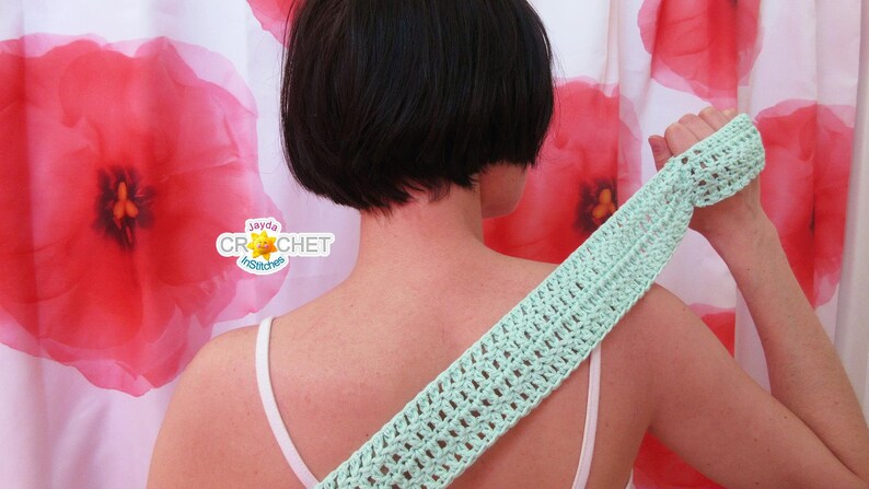 May include: A light green crocheted back scrubber with a textured pattern. The scrubber is being held by a person in front of a white shower curtain with red poppy flower designs.