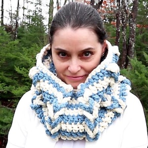 Peut inclure: Une femme porte une écharpe-cowl crochetée bleue et blanche. L'écharpe est épaisse et texturée, avec un motif répétitif. L'arrière-plan est une vue floue d'arbres et de feuillage verts. Le logo "Jayda Crochet in Stitches" est en haut à gauche.