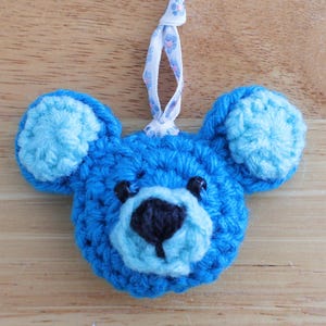 May include: A handmade blue crochet bear ornament with light blue ears and muzzle, and a black nose. The ornament has a white ribbon for hanging. The text "Jayda Crochet inStitches" is in the upper left corner.