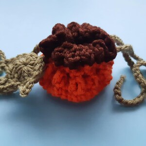 Puede incluir: Una bolsa de calabaza de ganchillo con la parte superior marrón y la inferior naranja. La bolsa tiene un cordón de ganchillo de color canela.
