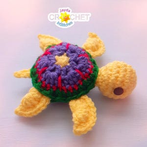 May include: A handmade crochet turtle toy. The turtle features a yellow head and flippers, a purple and green shell with red accents, and a brown eye. The image includes the text "Jayda Crochet InStitches".