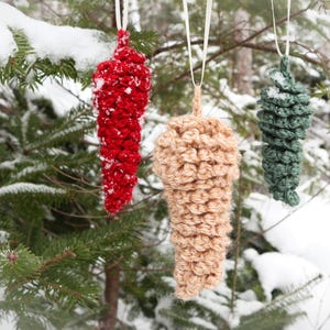May include: Three handmade crochet pinecone ornaments in red, tan, and green hang from a snow-covered evergreen tree. Each ornament is suspended by a white ribbon. The background is a blurred winter scene.