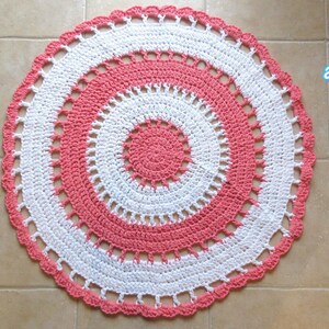 May include: A handmade, circular area rug in a crochet design. The rug features concentric circles of white and coral-pink, with a scalloped edge. The diameter is approximately 33.5 inches. Text on the image reads "Area Rug" and "Jayda Crochet inStitches".