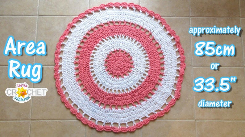 May include: A handmade, circular area rug in a crochet design. The rug features concentric circles of white and coral-pink, with a scalloped edge. The diameter is approximately 33.5 inches. Text on the image reads "Area Rug" and "Jayda Crochet inStitches".