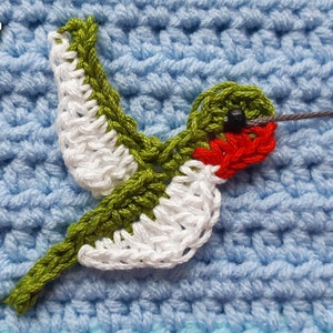 Puede incluir: Una aplicación de colibrí de ganchillo en verde, blanco y rojo con una cuenta negra para el ojo. El colibrí está volando con las alas extendidas. La aplicación está sobre un fondo de ganchillo azul claro.