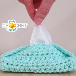 May include: A crocheted tissue box cover in a light turquoise color. The cover is rectangular with a textured stitch pattern and a folded edge. The text "Jayda Crochet inStitches" is in the upper left corner.