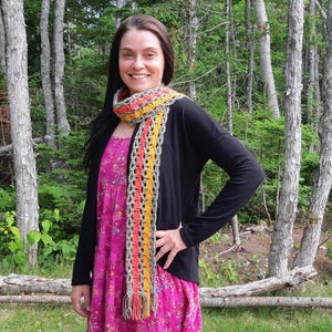 May include: A woman wearing a black cardigan over a pink floral dress, with a colorful crochet scarf. The scarf features orange, yellow, and gray sections. The background includes trees and green foliage, with the text "Jayda Crochet in Stitches" in the upper left corner.