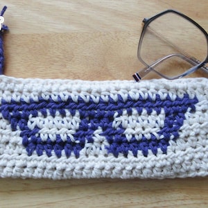 May include: White crocheted glasses case with a blue sunglasses design. The case has a purple drawstring.