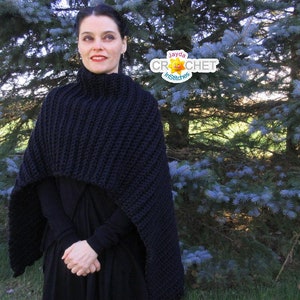 Può includere: Un poncho in crochet nero con collo alto e un design lungo e fluido. Il poncho è indossato da una persona in piedi davanti a un abete blu.