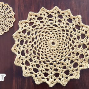 May include: Two beige crocheted doilies on a dark wood surface. One is small and round, the other is large with a starburst edge. The crochet pattern is open and airy, with a spiral center. The doilies are handmade and decorative.