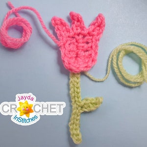 May include: A pink crocheted flower with a green stem and leaf. The flower is made using single crochet stitches and the stem and leaf are made using double crochet stitches. The flower is attached to the stem with a slip stitch. The flower is surrounded by yarn.